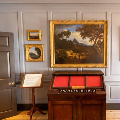 Photo of a musical instrument with a landscape oil painting behind it against a grey panelled wall.