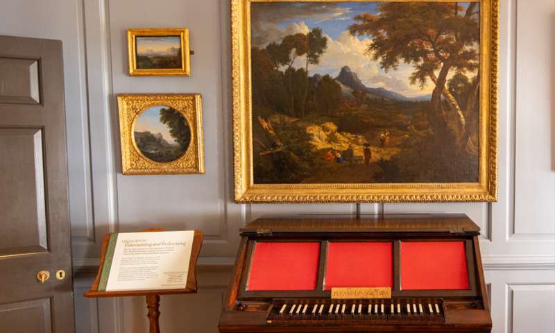 Photo of a musical instrument with a landscape oil painting behind it against a grey panelled wall.