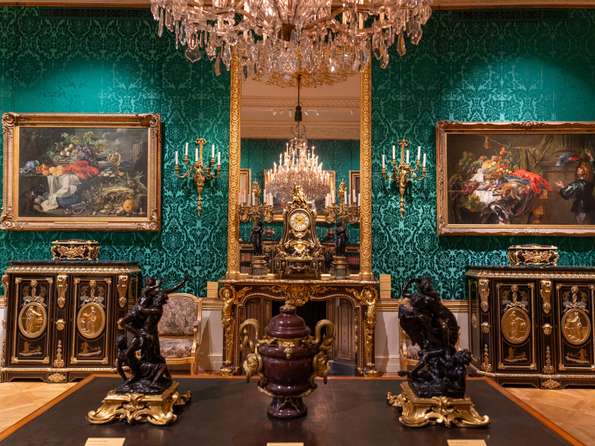 A fireplace set against green wallpaper, flanked by ornate cabinets. A huge chandelier is in view.