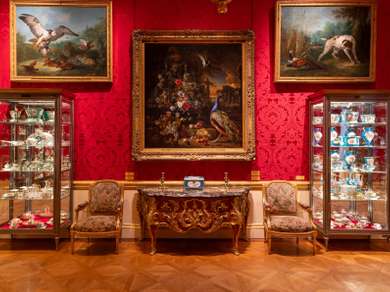 Three large classical paintings on a wall with bright red wallpaper, underneath there's an ornate table.