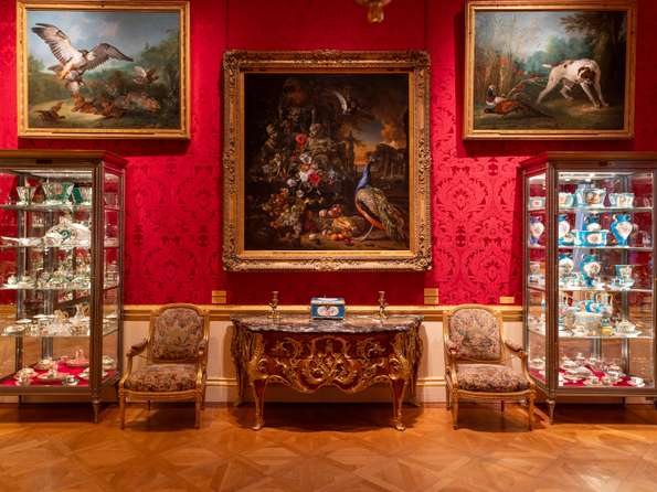 Three large classical paintings on a wall with bright red wallpaper, underneath there's an ornate table.